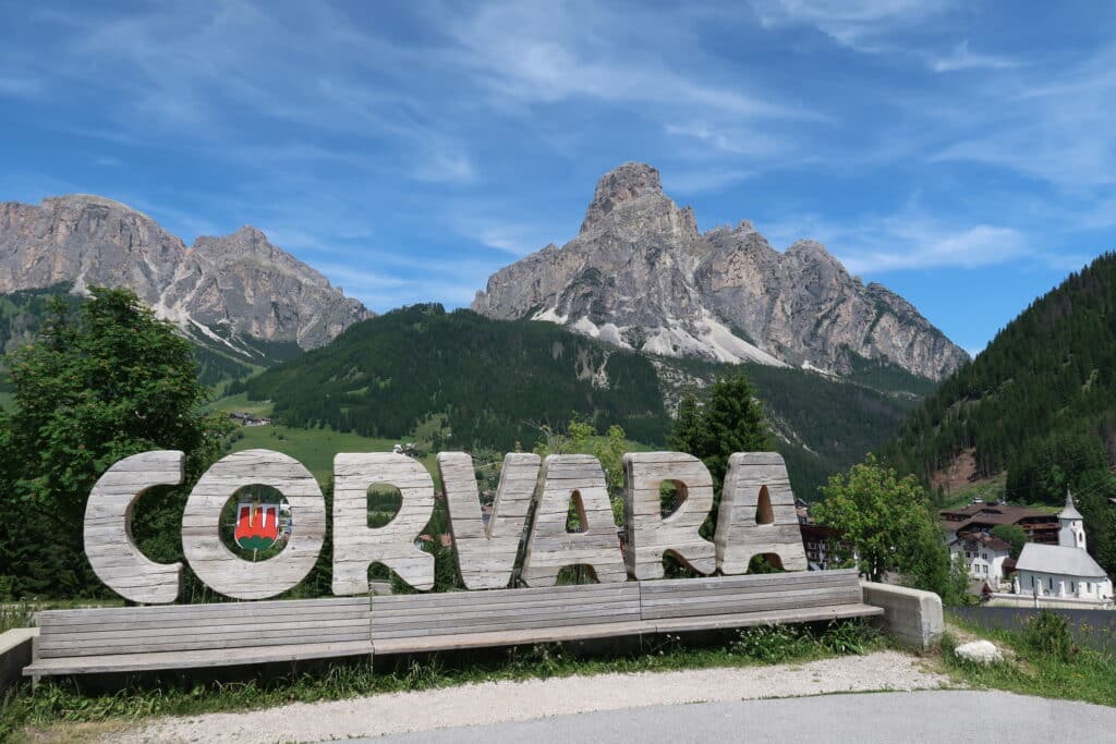 Large wooden Corvara town sign outdoors in front of the Italian alps on a sunny day